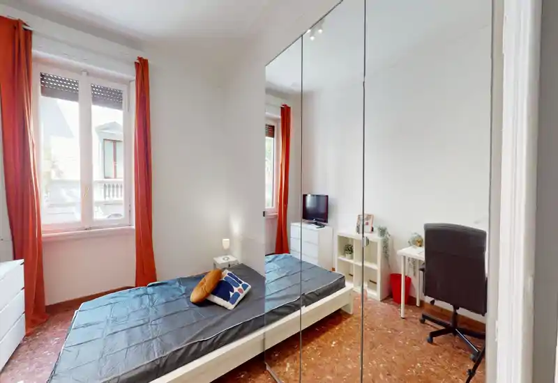 Bright, neatly staged bedroom with a double bed, bedside lamp and desk area; large window with orange curtains provides natural light and mirrored wardrobe adds depth.