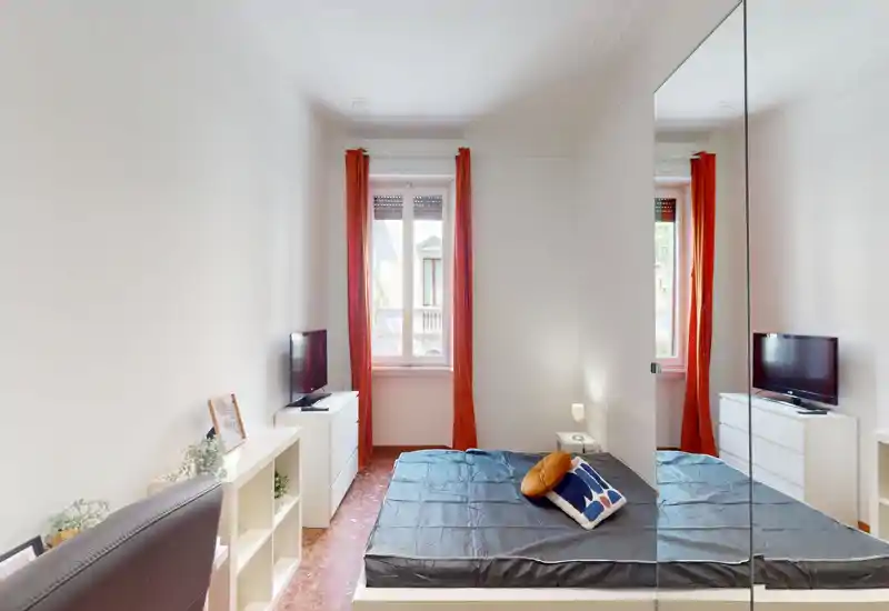 Bright, minimalist bedroom with a double bed, decorative pillows and large mirrored wardrobe creating extra depth. Natural light from a central window and warm orange curtains add character; small bedside table and TV provide practical comfort.