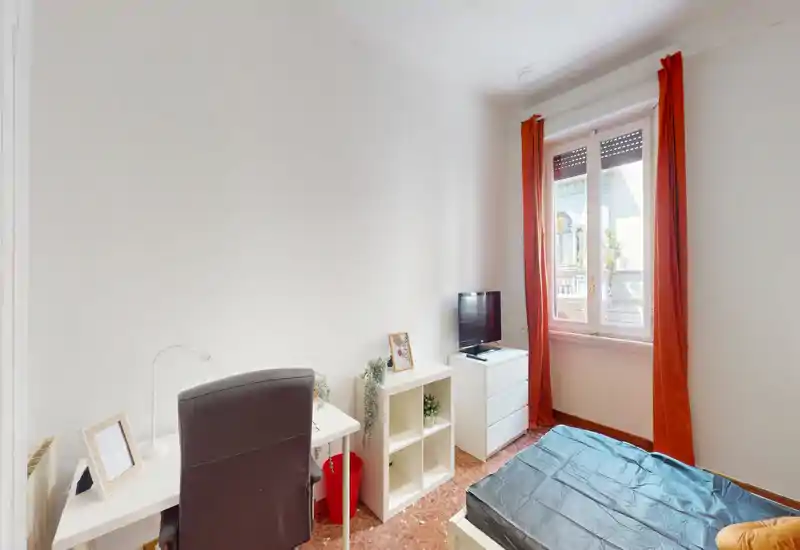 Bright single bedroom with a bed, desk and TV near a large window. Clean minimalist décor with warm terracotta floor and orange curtains providing a cozy accent.