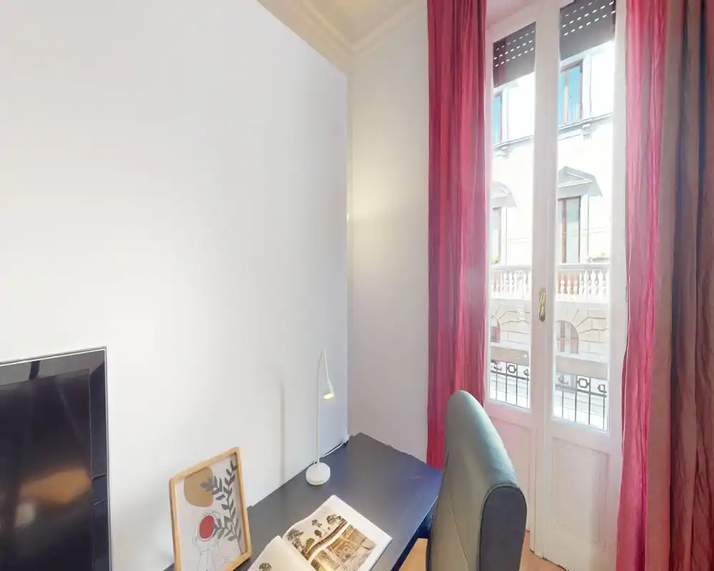 Bright corner of a living area with a desk, armchair, TV and a balcony door letting in natural light; simple, modern decor with red curtains and a small desk lamp.