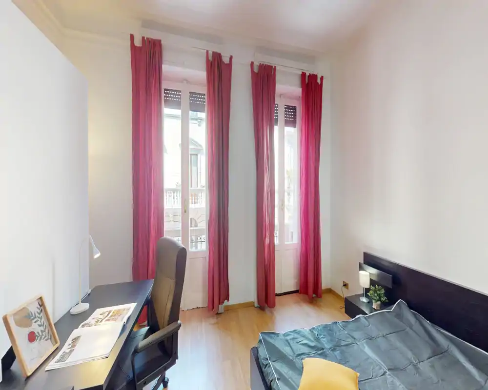 Bright, minimalist bedroom with tall windows dressed in deep red curtains, a double bed with simple headboard, bedside lamp and a desk with chair—ideal for work and rest.