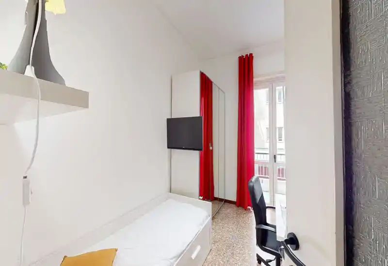 Camera da letto piccola e luminosa con letto singolo, armadio con anta a specchio, TV a parete e scrivania vicino alla finestra con tende rosse che filtrano la luce naturale.