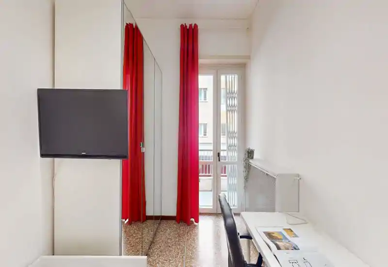 Camera compatta con luce naturale, tende rosse a tutta altezza, TV a parete e una piccola scrivania vicino alla finestra — pulita, essenziale e funzionale.