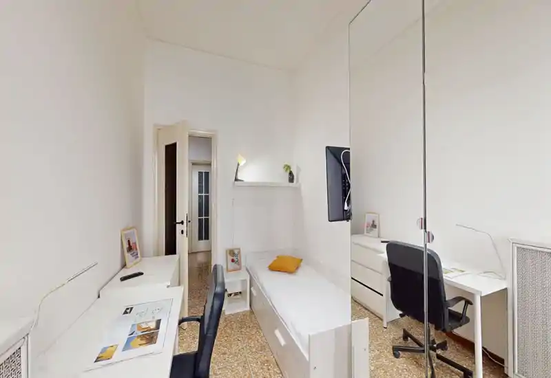 Camera singola luminosa dallo stile pulito e minimale: letto singolo, due scrivanie con sedie, mensola a parete e dettagli decorativi semplici. Ideale per uno studente o un professionista che cerca spazio compatto e ben illuminato.