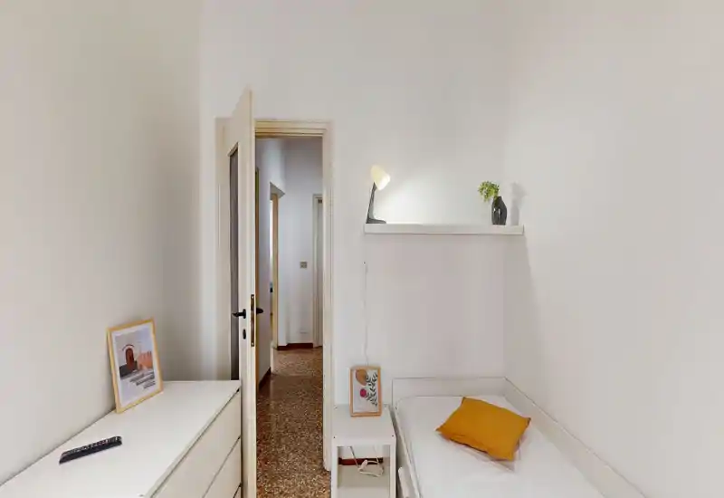 Camera singola compatta con letto singolo ben rifatto, mensola con lampada e vaso e piccolo comodino. Pareti bianche e arredo minimale creano uno spazio luminoso e ordinato.