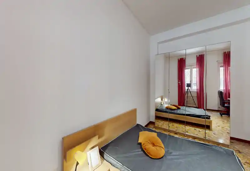 Dormitorio luminoso y ordenado con cabecero de madera, ropa de cama oscura y un cojín redondo como acento. El armario con espejo y la luz natural amplían visualmente la habitación.