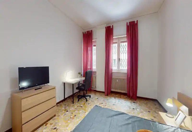 Dormitorio luminoso y amueblado de forma sencilla: esquina de la cama visible, mesita con lámpara, cómoda con TV y una zona de trabajo junto a las ventanas con cortinas rojas que aportan luz natural.