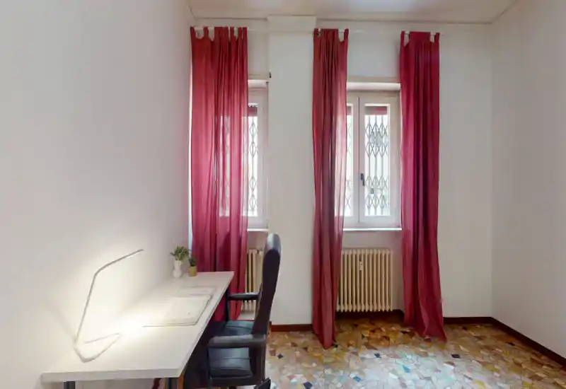 Habitación pequeña y luminosa acondicionada como zona de trabajo/salón con escritorio, silla de oficina y dos ventanas altas con cortinas rojas que dejan entrar luz natural.