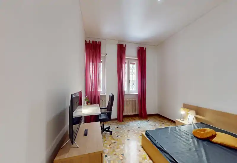Dormitorio luminoso con cama doble, lámpara de mesita, escritorio y TV; cortinas rojas en dos ventanas y suelo de terrazo.