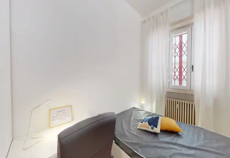 Dormitorio compacto y ordenado con una cama individual, cojines decorativos y luz natural suave desde la ventana. Una pequeña lámpara de escritorio y una decoración neutra crean un espacio funcional y tranquilo.