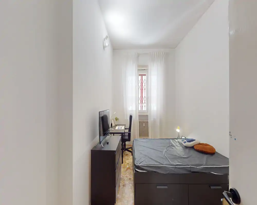 Dormitorio luminoso y de mobiliario sencillo con cama con almacenaje, TV sobre cómoda y un pequeño escritorio junto a la ventana que deja entrar luz natural a través de cortinas traslúcidas.