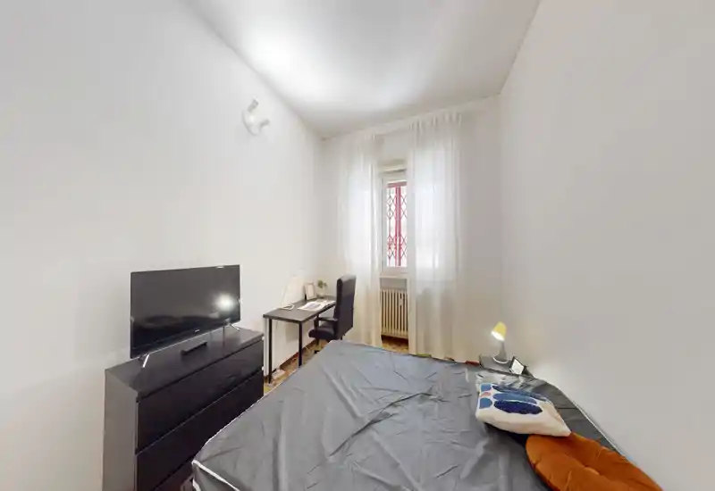 Habitación compacta y luminosa con cama doble, lámpara de noche, televisor sobre cómoda y un pequeño escritorio junto a la ventana. Estilo limpio y luz natural suave, ideal para estancias cortas o estudiantes.