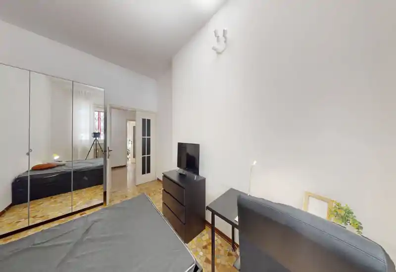 Dormitorio moderno y luminoso con cama doble, armario con puertas espejadas y un pequeño escritorio con TV. Suelo tipo parquet, luz natural agradable y mobiliario minimalista perfecto para un anuncio de alquiler.