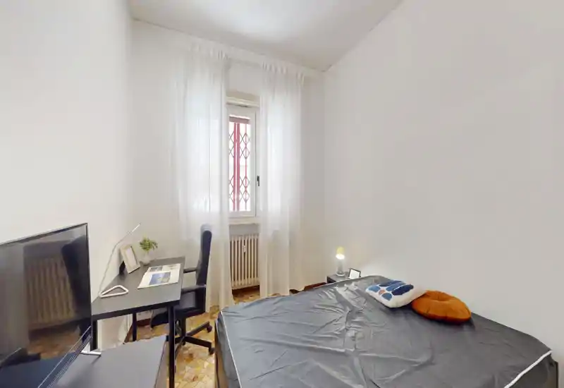 Dormitorio luminoso y minimalista con cama doble, lámpara de noche y un pequeño escritorio junto a la ventana — perfecto para dormir y trabajar. Las cortinas translúcidas dejan pasar la luz natural conservando la privacidad.