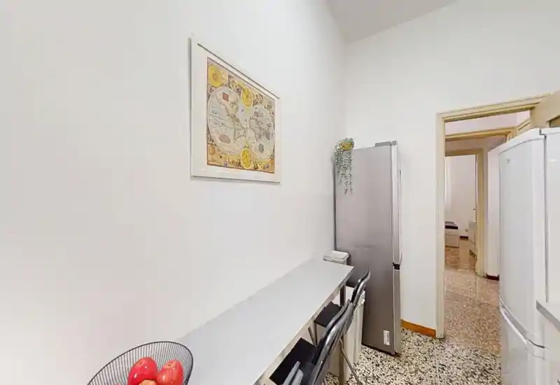 Cocina estrecha tipo pasillo con encimera delgada, frigorífico alto y sillas; diseño sencillo y funcional con cuadro decorativo y un cuenco de fruta.