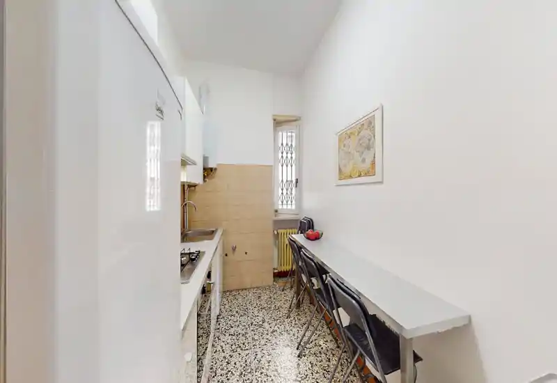 Cocina en galería estrecha con muebles blancos, placa y fregadero a la izquierda y una barra larga con taburetes a la derecha; luminosa, bien cuidada y dispuesta de forma eficiente para espacios urbanos compactos.