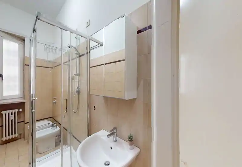Baño compacto con mampara de ducha de cristal, mueble con espejo, lavabo y bidé; azulejos beige neutro y ventana esmerilada que aporta luz natural.