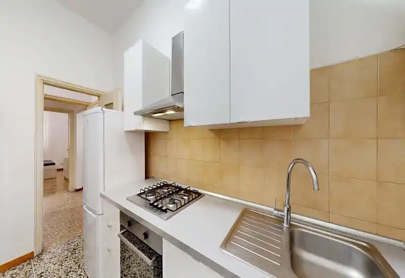 Cocina compacta y funcional con vitrocerámica a gas de cuatro fuegos, horno integrado, fregadero de acero inoxidable y armarios altos; salpicadero de azulejos neutros y encimera luminosa, ideal para el uso diario.