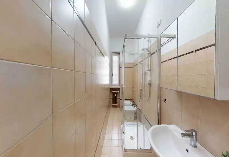 Baño estrecho alicatado con ducha con mampara de vidrio, lavabo mural y mueble con espejo; espacio luminoso, limpio y funcional ideal para un piso compacto.