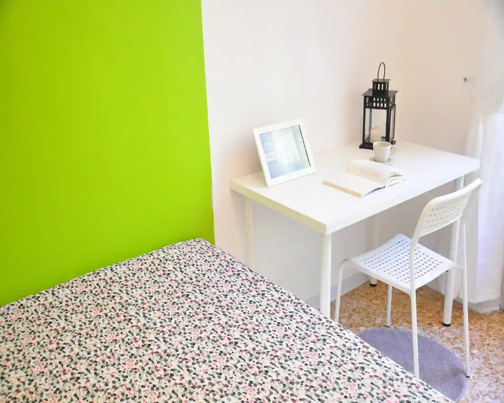 Chambre lumineuse et soignée avec un lit à motif au premier plan et un bureau blanc près de la fenêtre ; mur d'accent vert et déco minimaliste pour une atmosphère moderne et aérée.