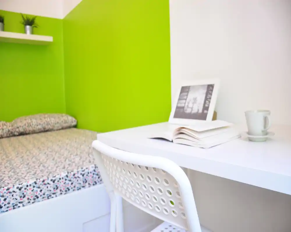 Coin chambre lumineux et compact avec un lit simple à motif, bureau et chaise blancs. Le mur vert et les petites plantes apportent une touche fraîche et moderne — idéal pour une location cosy.