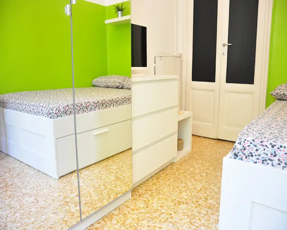 Chambre lumineuse et moderne avec deux lits blancs, linge de lit à motifs et murs vert vif. L'espace est propre, bien éclairé et aménagé pour une ambiance fraîche et jeune.