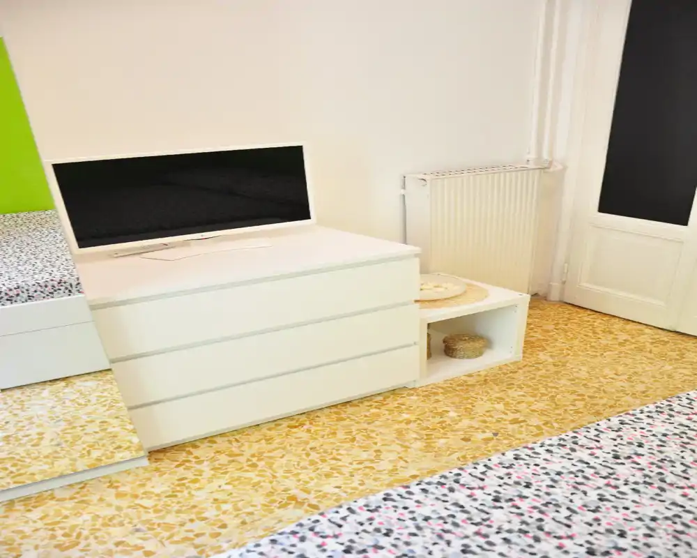 Coin de chambre lumineux et minimaliste avec commode blanche, télévision et partie du matelas visible. Style propre et moderne, sol en terrazzo apportant une ambiance fraîche et dégagée.