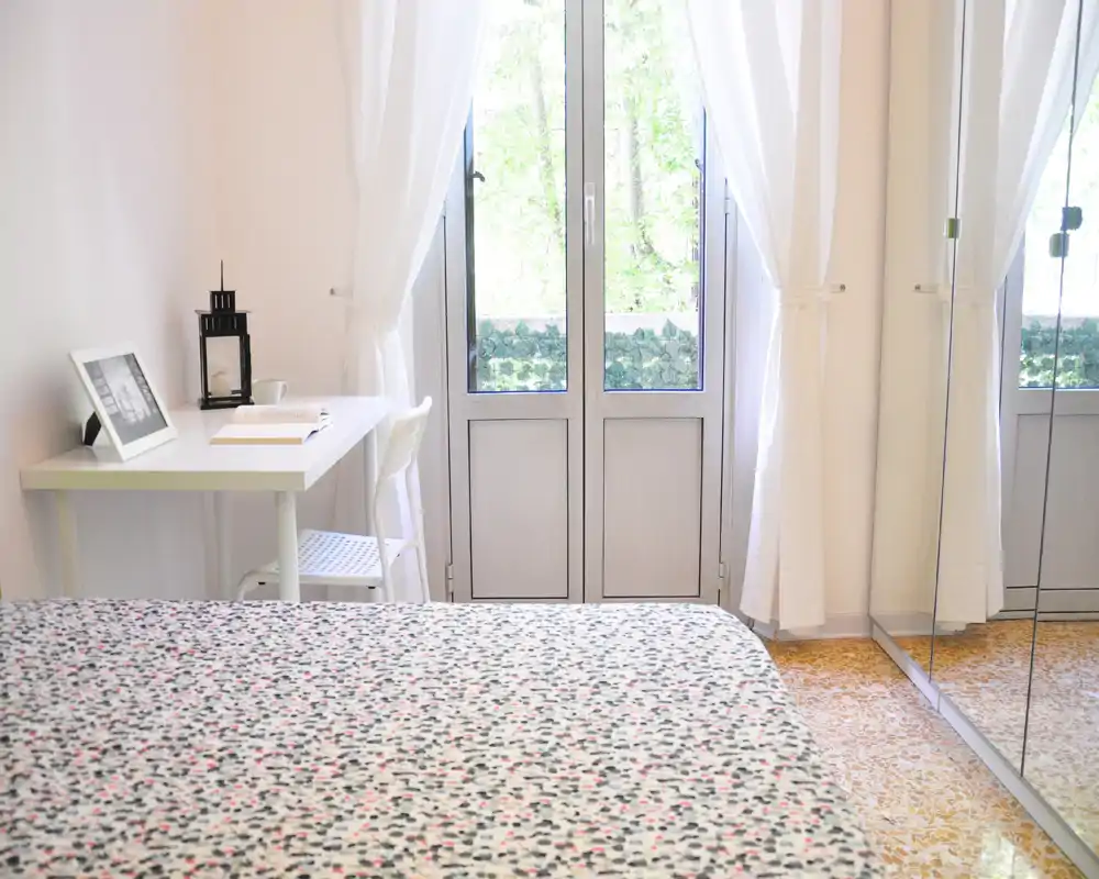 Chambre lumineuse et aérée avec une couette à motifs, des voilages blancs et un petit bureau près de la fenêtre. La garde-robe miroir apporte de la profondeur.