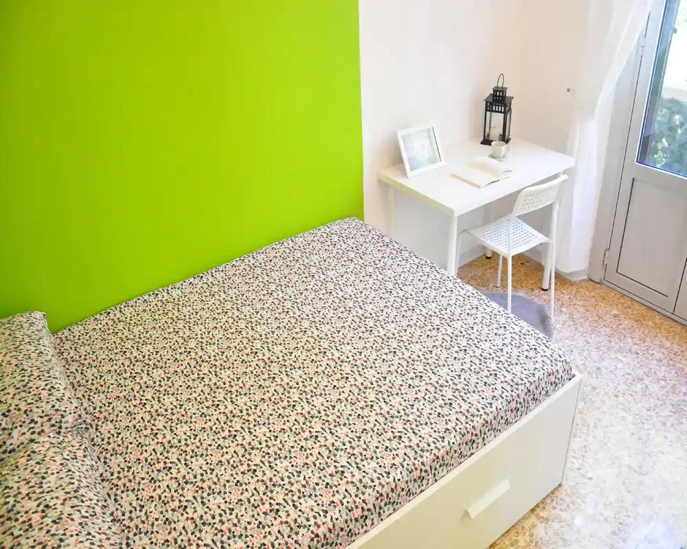 Chambre moderne et lumineuse avec un grand lit double occupant la plupart de l'espace, une literie à motifs et un petit bureau blanc près de la fenêtre. Le mur vert en accent et la lumière naturelle donnent une ambiance fraîche et accueillante, idéale pour une annonce locative.