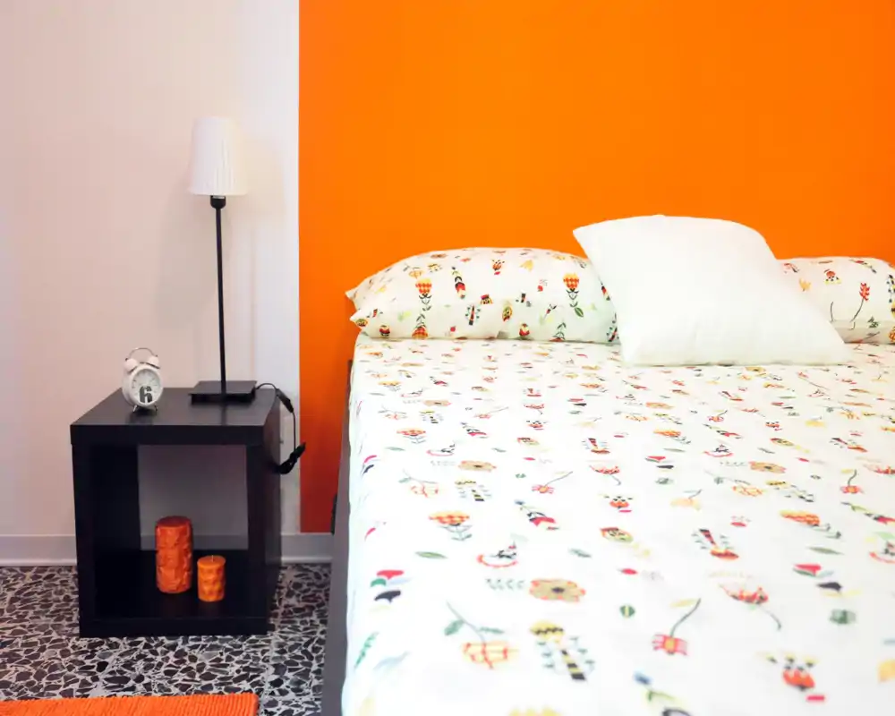 Camera da letto luminosa e vivace con biancheria a fantasia e parete accentuata arancione. Il letto occupa la scena e c'è un comodino minimale con lampada e orologio.