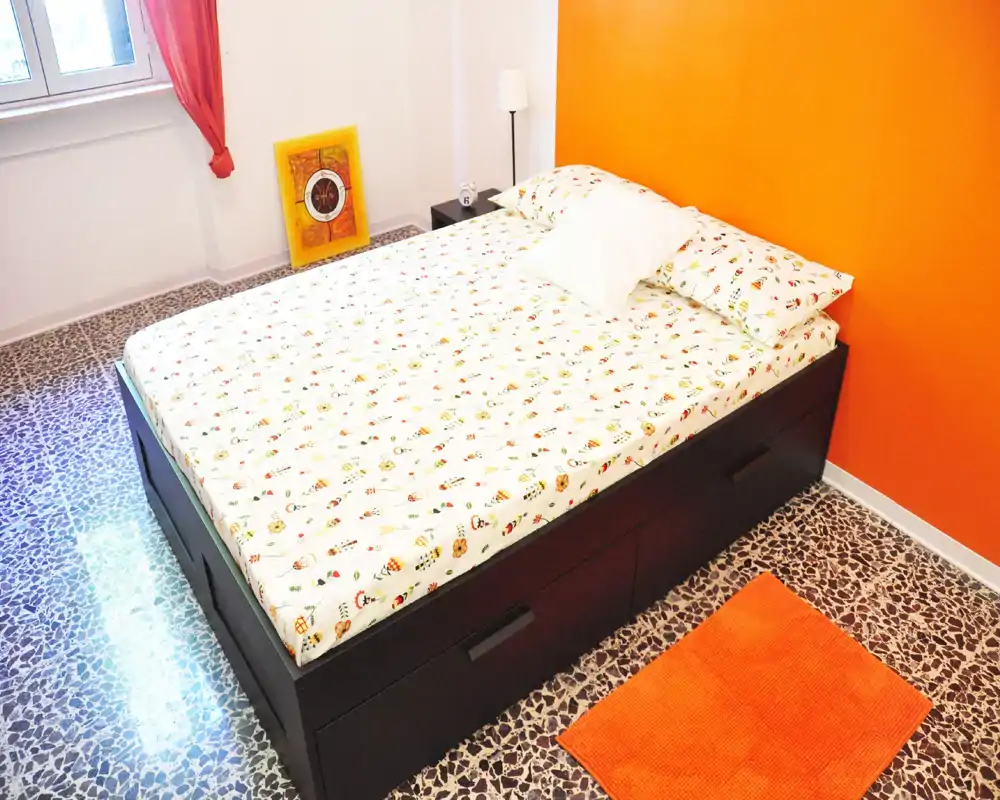 Camera da letto luminosa e ben composta con un grande letto contenitore in legno scuro, biancheria a fantasia e una calda parete arancione; la luce naturale dalla finestra e la lampada rendono l'ambiente accogliente.