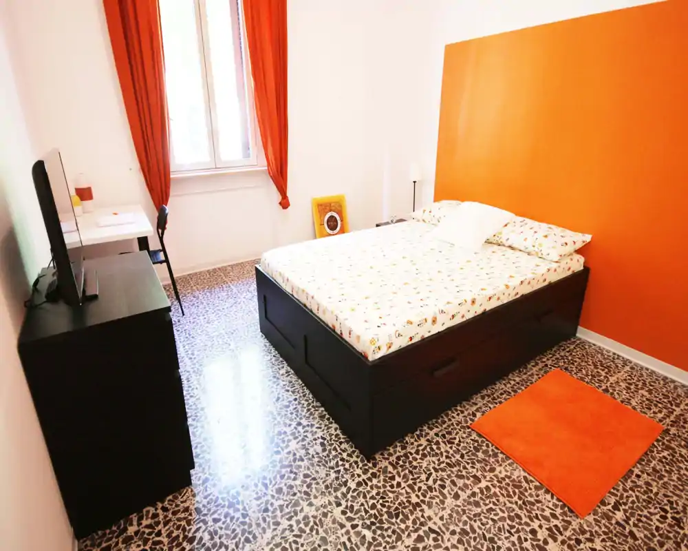 Camera da letto luminosa con accenti arancioni, letto matrimoniale, scrivania e TV; pavimento in graniglia e tende calde creano un ambiente accogliente e moderno.