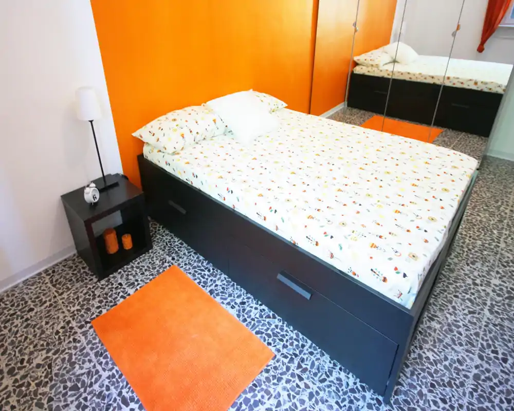 Camera da letto moderna e luminosa con letto matrimoniale, armadio con specchi e parete d'accento arancione; ambiente ben illuminato e arredato con cura.