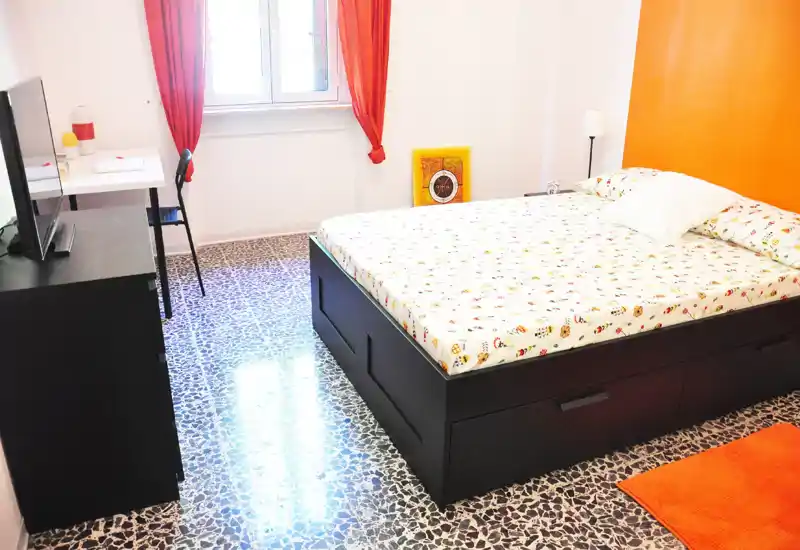 Camera da letto luminosa e ben allestita con un grande letto contenitore nero, biancheria a fantasia e accenti arancioni caldi. La luce naturale dalla finestra valorizza il pavimento in graniglia e l'angolo studio ordinato.