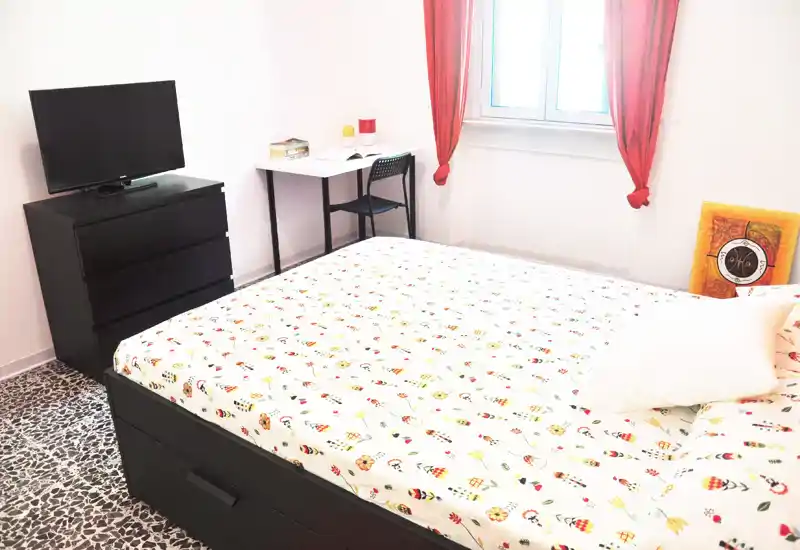 Camera da letto luminosa e ordinata con un ampio letto con biancheria a fantasia, comò con TV e una piccola scrivania vicino alla finestra — ideale per brevi soggiorni o studenti.