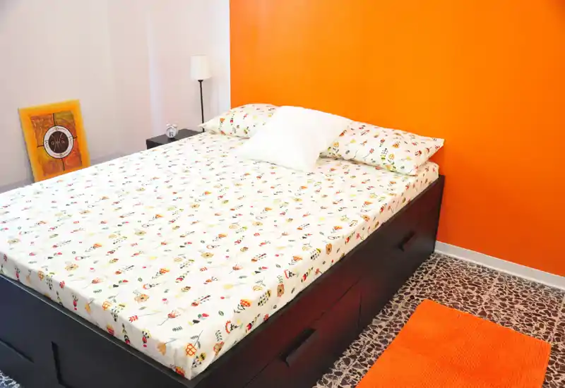 Camera da letto luminosa e moderna con un grande letto e biancheria fantasia, parete arancione a contrasto e comodi cassetti sotto la struttura: ambiente accogliente e ben illuminato.