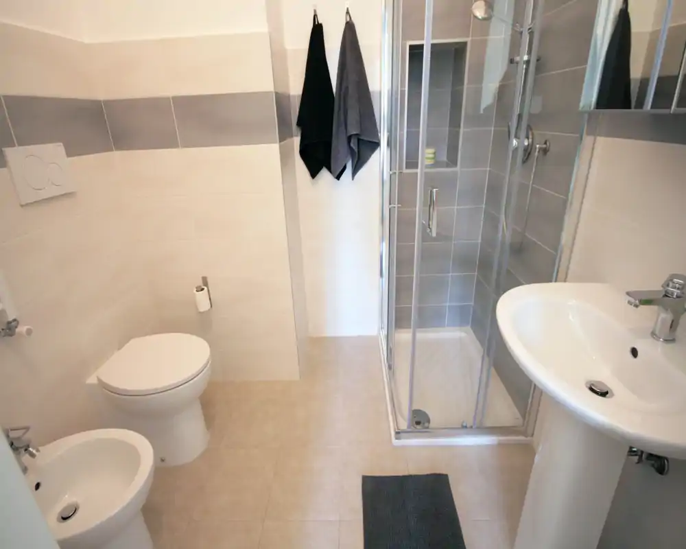 Bagno moderno e luminoso con cabina doccia, WC, bidet e lavabo a colonna; piastrelle neutre e asciugamani appesi per un aspetto pulito e contemporaneo.