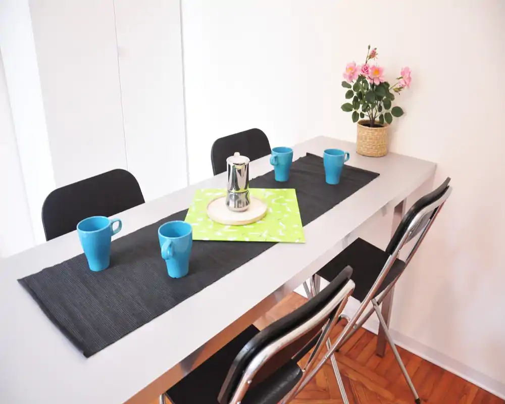 Zona pranzo moderna e luminosa con un tavolo allungato apparecchiato per quattro, tazze blu, una caffettiera e fiori decorativi — perfetta per pasti informali.