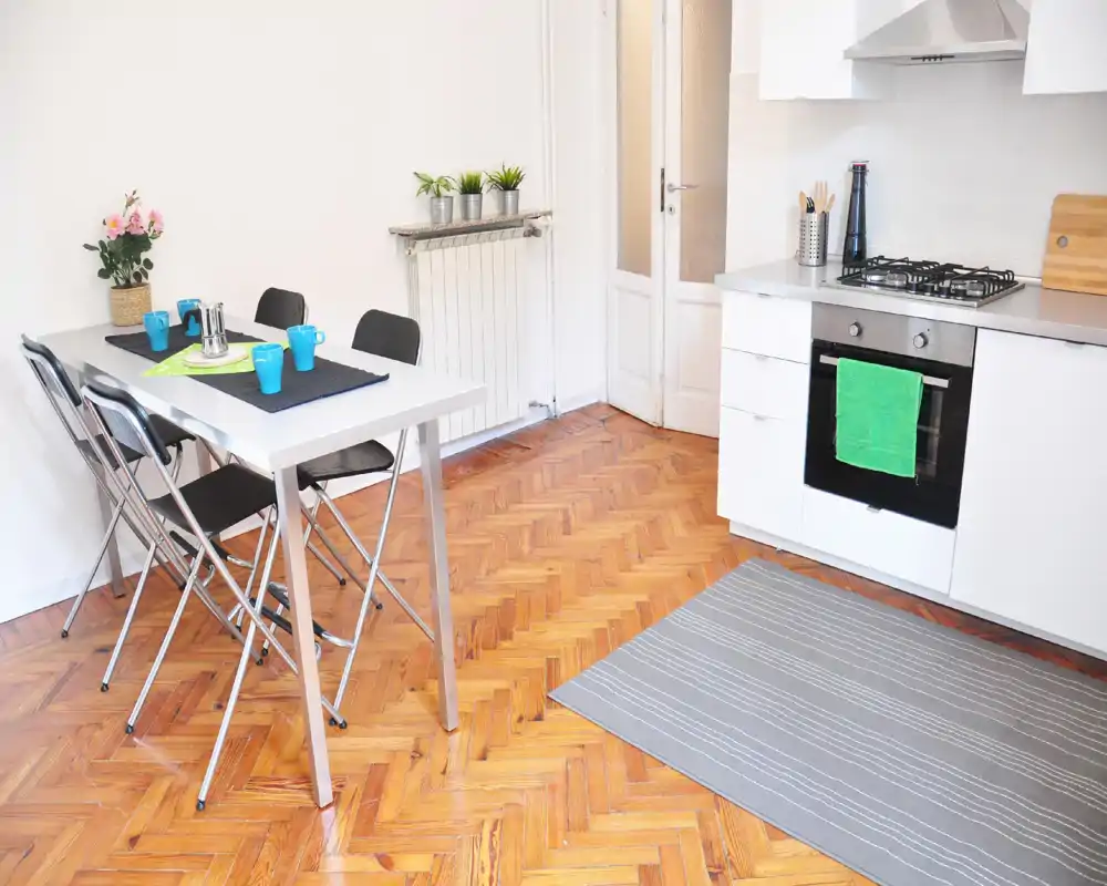 Cucina moderna e luminosa con mobili bianchi, forno incassato e un piccolo tavolo da pranzo per quattro; pavimento in parquet a spina di pesce e piante decorative rendono l'ambiente accogliente.