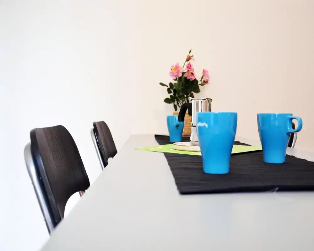 Allestimento da pranzo luminoso e moderno con tovaglietta semplice, tazze blu e un piccolo centrotavola floreale — perfetto per colazioni o pasti informali.