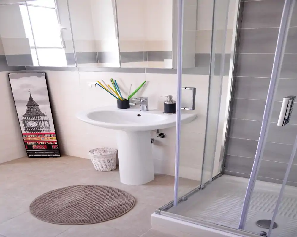 Bagno moderno e luminoso con lavabo a colonna, specchiera e box doccia in vetro. Piastrelle neutre e accessori semplici creano un aspetto pulito e contemporaneo.