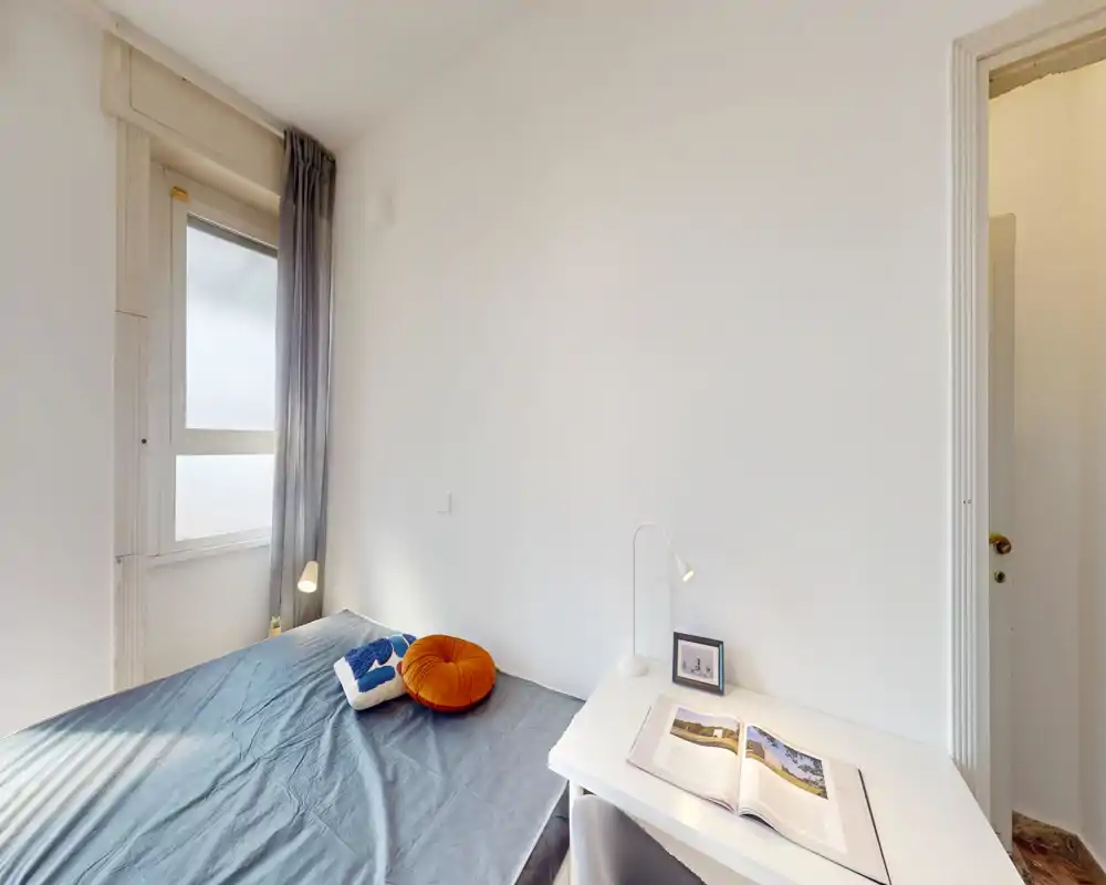 Small, bright bedroom with a double bed set against a white wall, a window providing soft natural light, and a tidy desk with a reading lamp — ideal for a single occupant or couple.