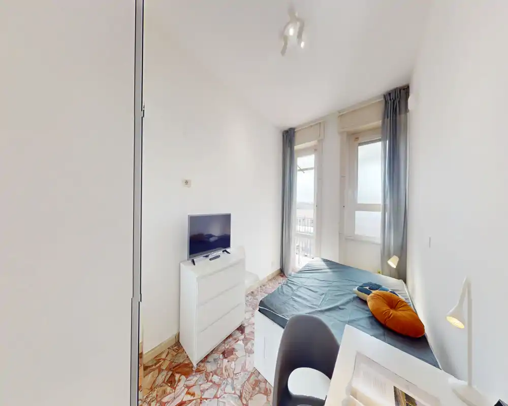 Bright single bedroom with a neatly made bed, bedside lamp and a chest of drawers with a TV. Large window/door provides natural light and a small balcony view, ideal for a compact rental.