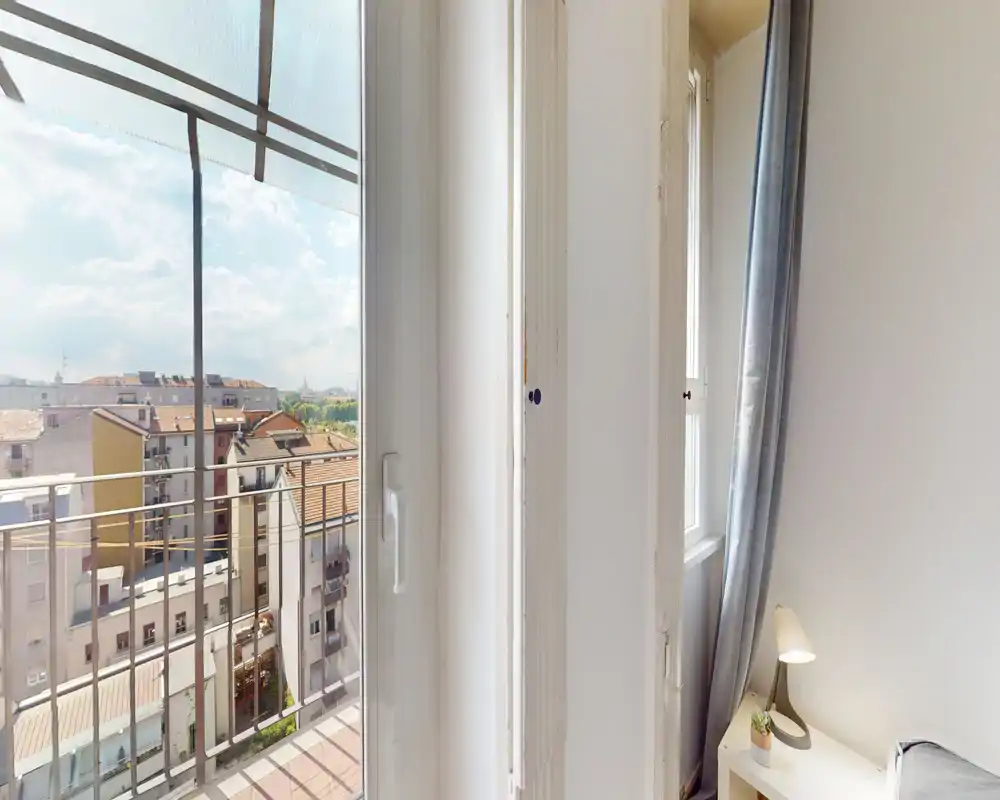 Small bright bedroom corner with a balcony view: a bedside table with lamp and a partially visible bed at the frame edge, plus a large glass door opening to a sunny balcony and city view.
