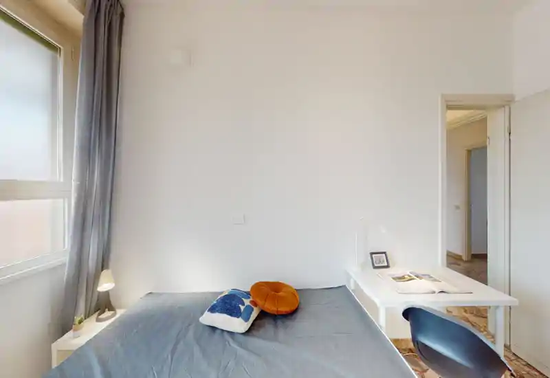 Chambre lumineuse et minimaliste avec un lit double, une lampe de chevet et un petit bureau près du lit. La lumière naturelle entre par une grande fenêtre et les coussins ajoutent une touche chaleureuse.