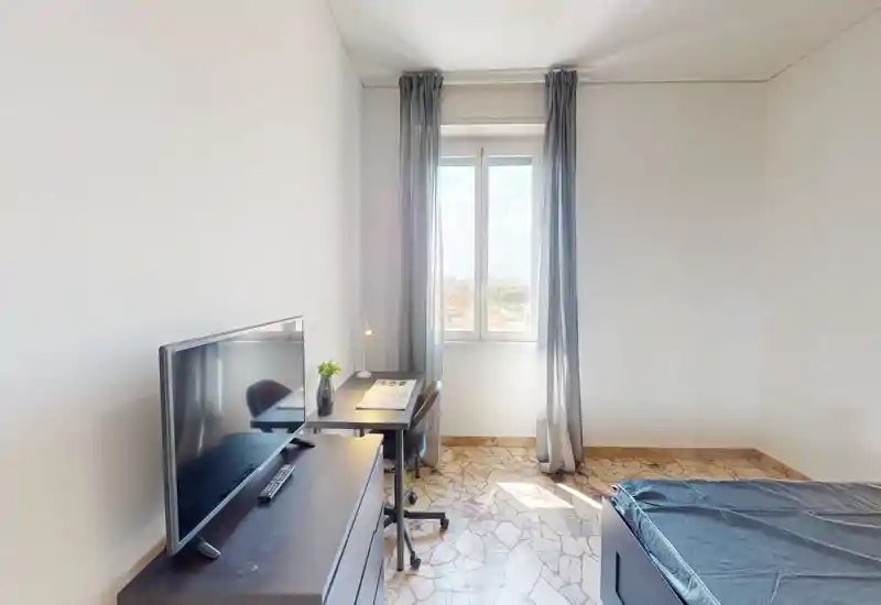 Chambre lumineuse et sobre avec lit simple, bureau et TV ; grande fenêtre apportant une belle lumière naturelle et un style minimaliste.