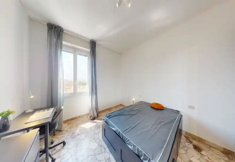 Chambre lumineuse et minimaliste avec lit double, lampes de chevet et un petit bureau près de la fenêtre. La lumière naturelle et les tons neutres lui confèrent une atmosphère aérée et fonctionnelle.