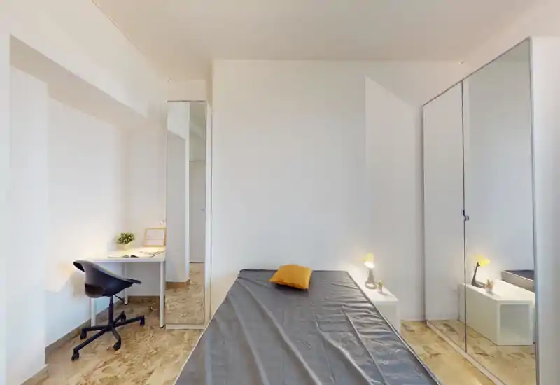 Minimal, well-lit single bedroom with a centrally framed bed, mirrored wardrobe and a compact desk — clean, modern and functional for a short-term stay.