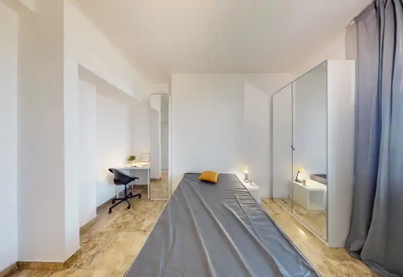 Minimal, well-lit bedroom with a single bed centered in the frame, a bedside table with a lamp and a mirrored wardrobe that adds depth. A small work desk and chair sit to the left; neutral tones and polished floor create a clean, modern feel.