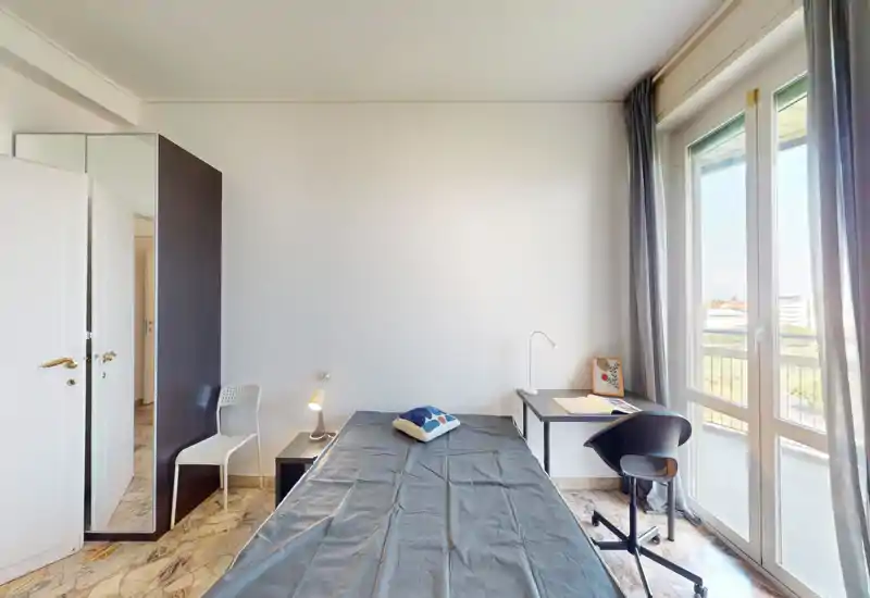 Camera da letto luminosa e arredata in modo semplice con un letto singolo, comodino e scrivania accanto a una grande finestra che offre molta luce naturale.